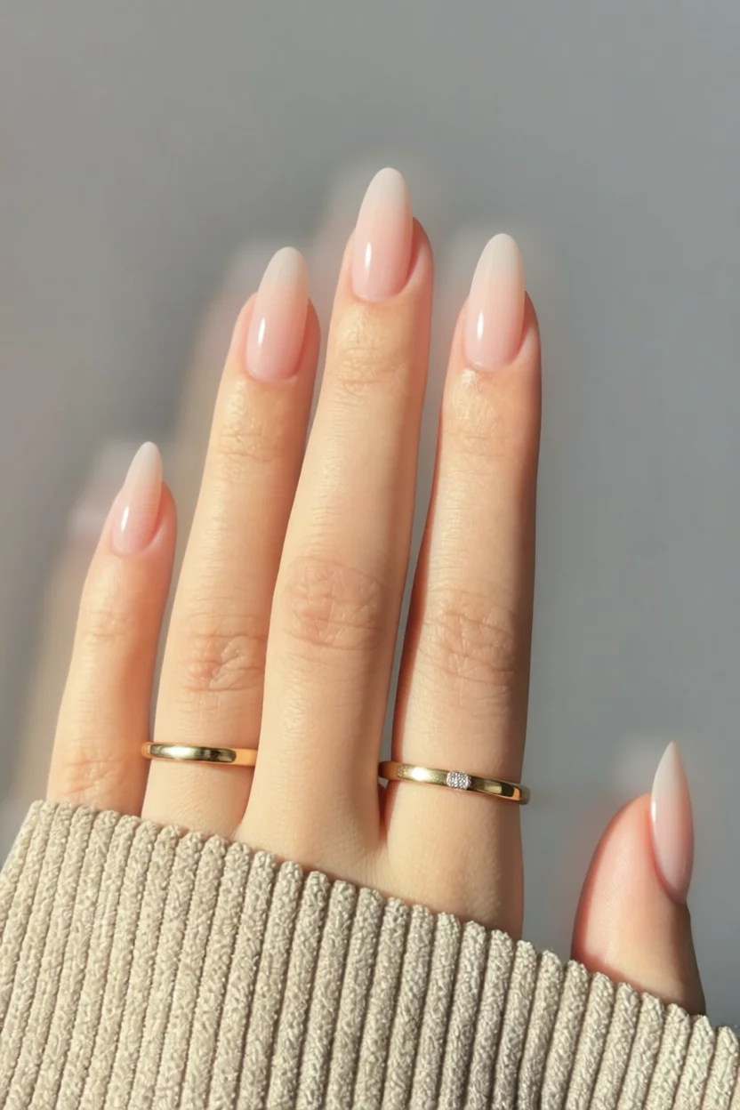 Mano con uñas largas en forma almendrada color nude rosado translúcido con acabado brillante tipo cristal y anillos dorados