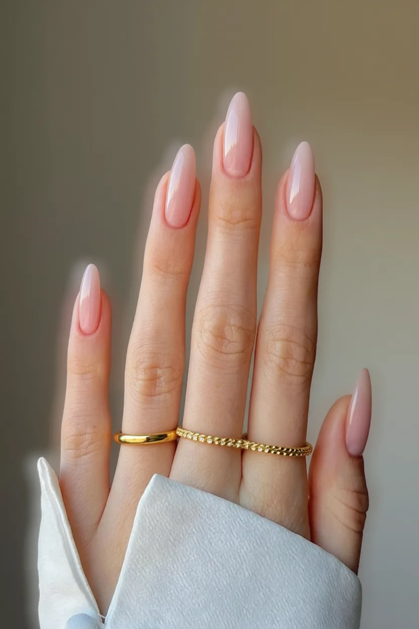Mano con uñas largas forma almendra en tono rosa nude brillante y dos anillos dorados en el dedo anular