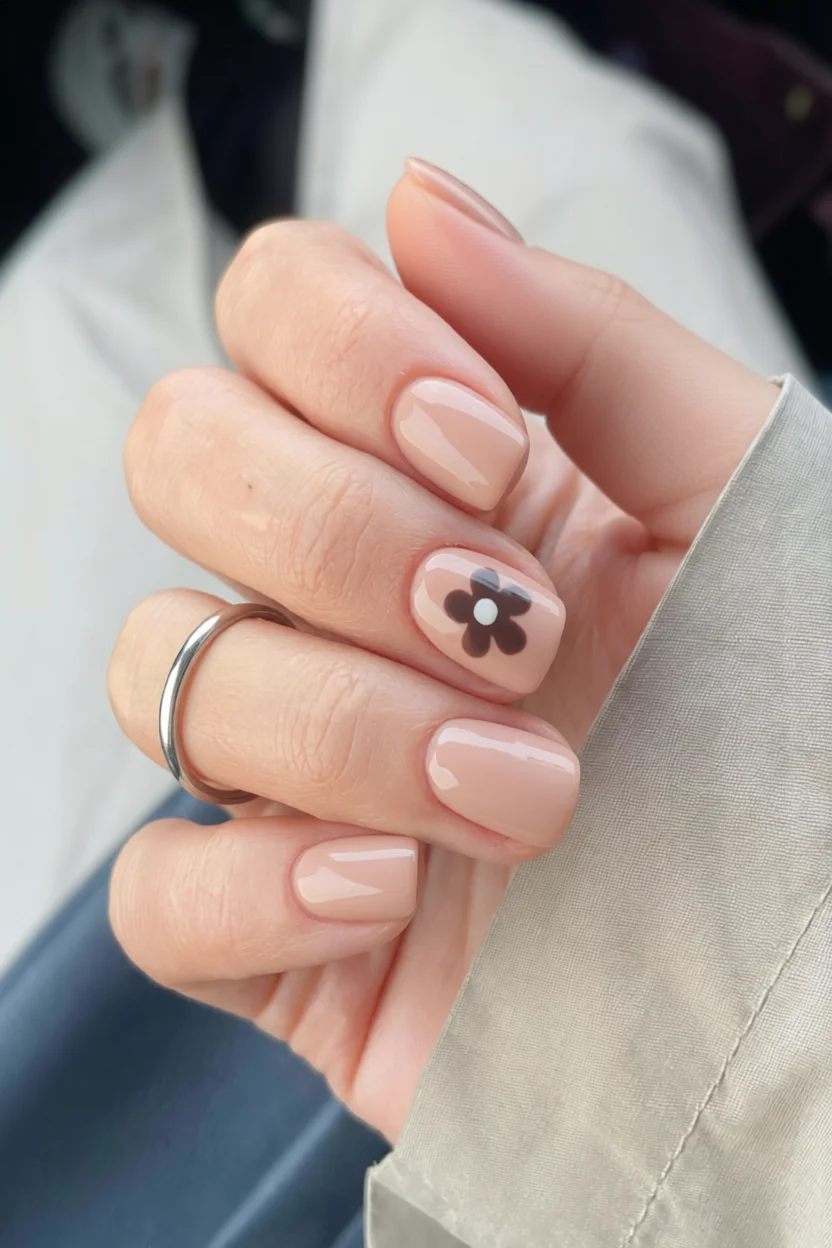 Manicura corta en tono nude con una uña decorada con pequeña flor color chocolate y punto central blanco