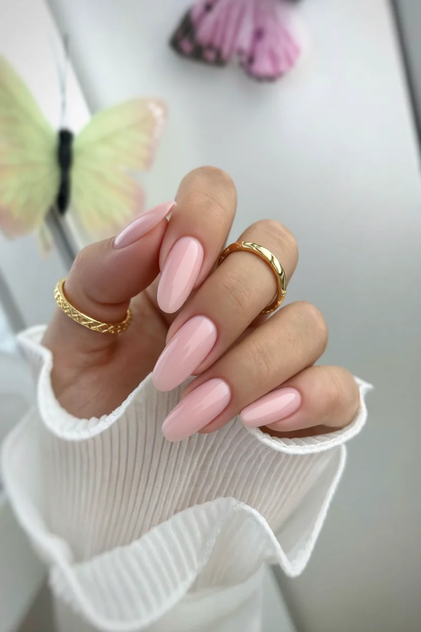 Mano con uñas rosa nude en forma almendra y anillos dorados, mariposas decorativas al fondo