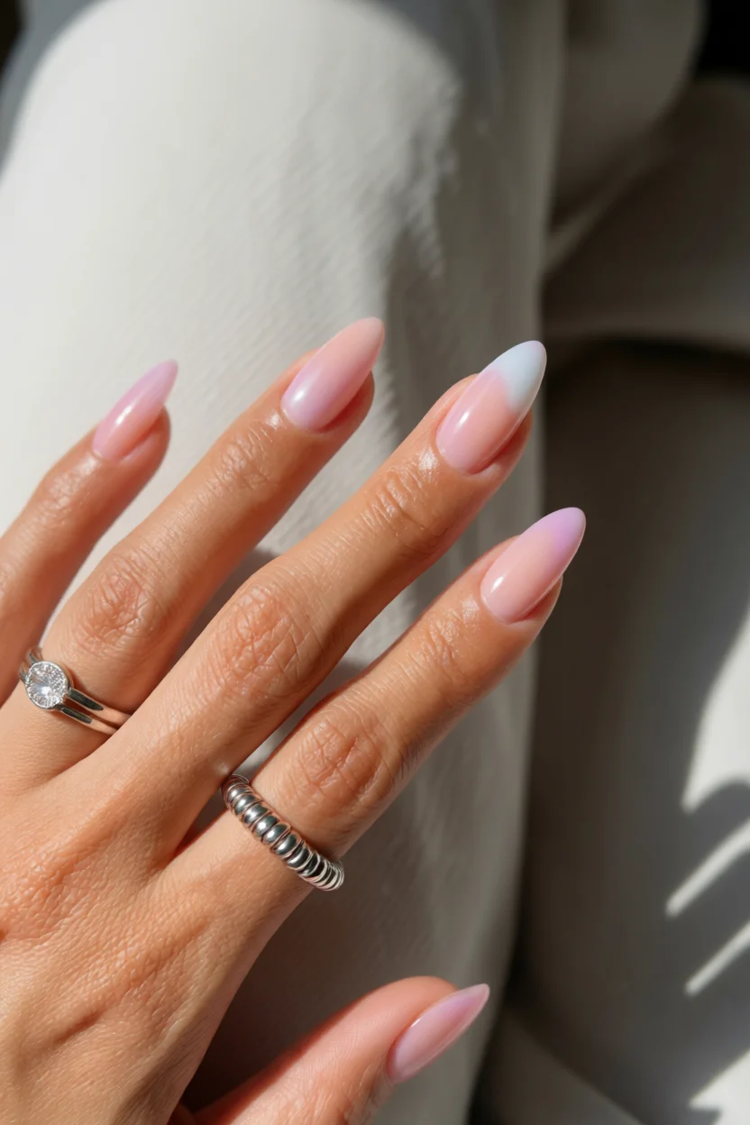 Mano con uñas ovaladas largas en degradado rosa pastel a blanco con brillo gel y anillos decorativos