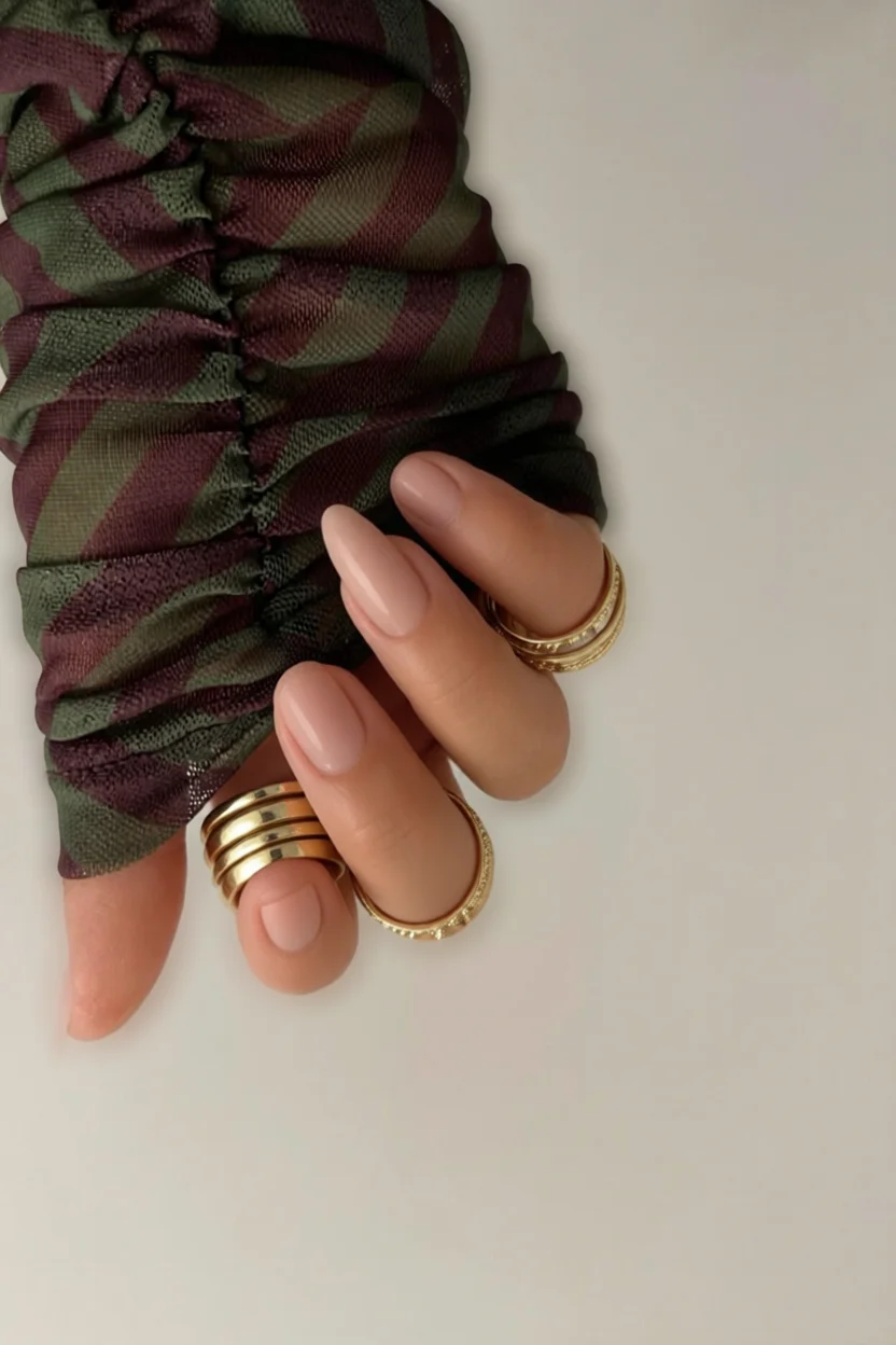 Mano con uñas de gel largas forma almendra color nude con anillos dorados sobre fondo de tela estampada