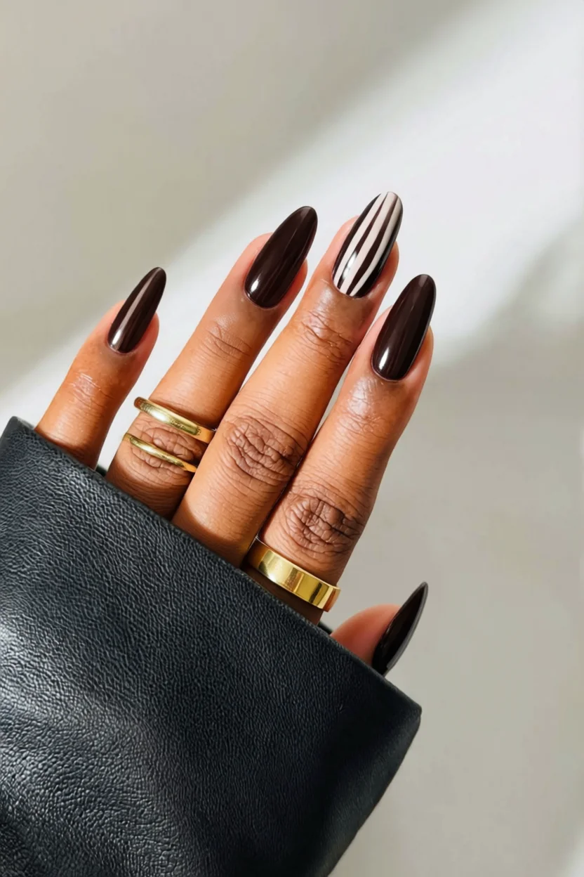 Mano con uñas largas almendradas color chocolate intenso y una con franjas blancas, anillos dorados