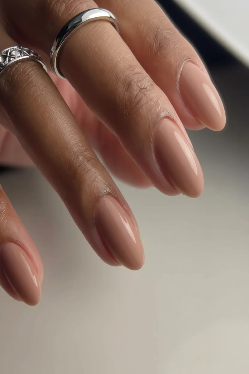 Mano con uñas barnizadas en tono marrón nude brillante y forma almendrada, con anillos plateados