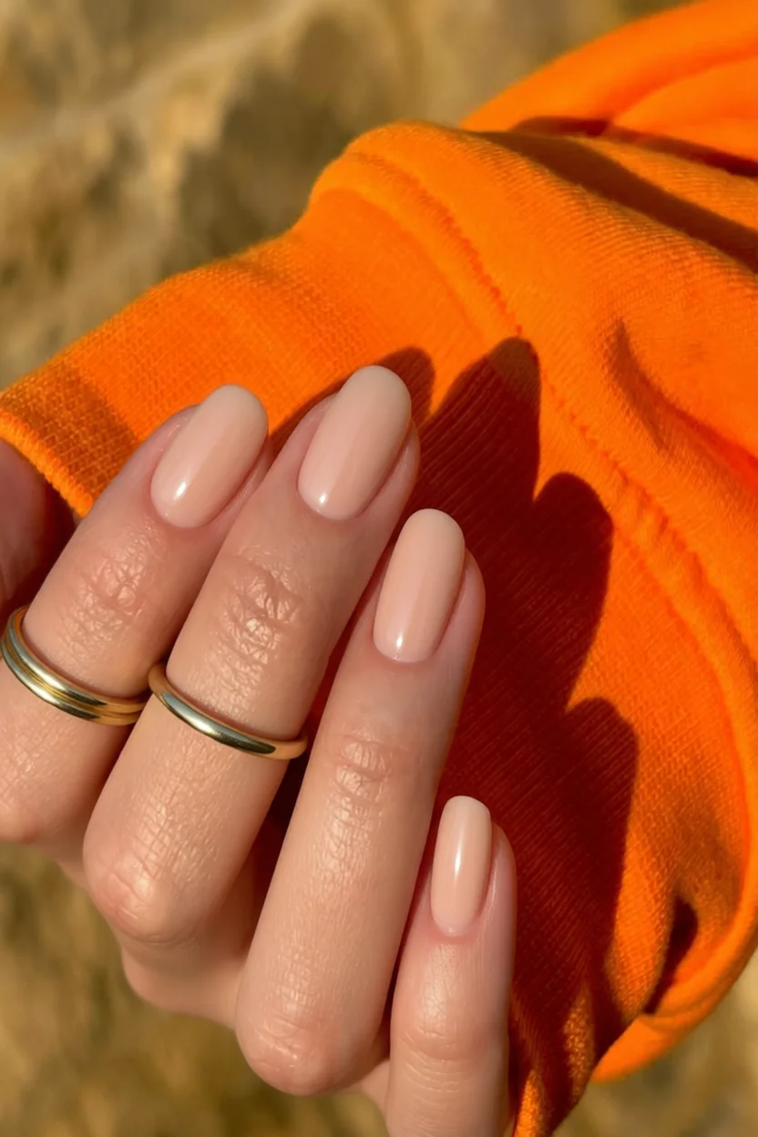 Mano con uñas ovaladas en tono nude y esmalte brillante sobre fondo naranja