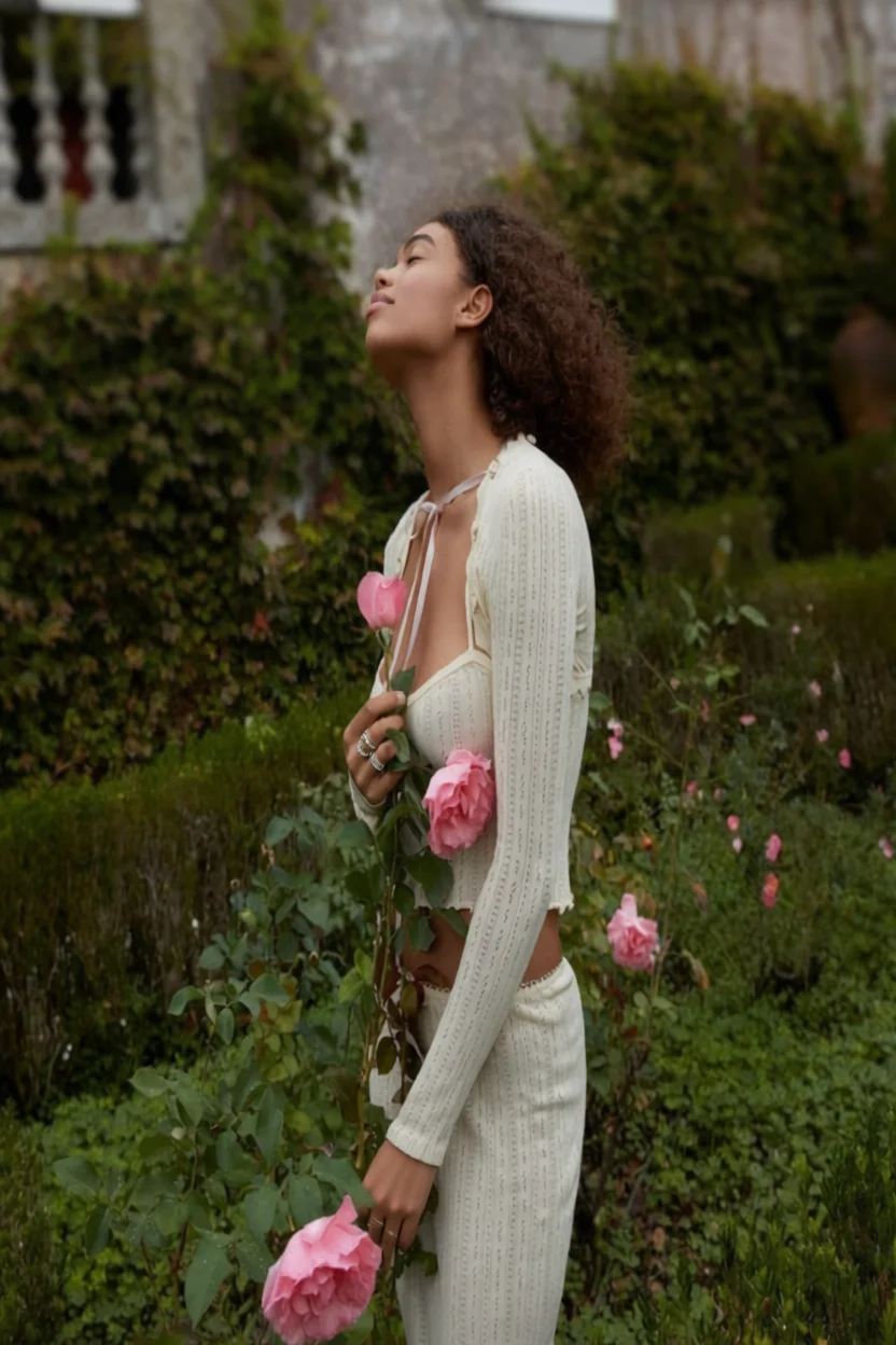 Mujer de piel morena con cabello rizado, vestida con conjunto blanco crema, sosteniendo rosas rosadas en un jardín con los ojos cerrados en actitud serena