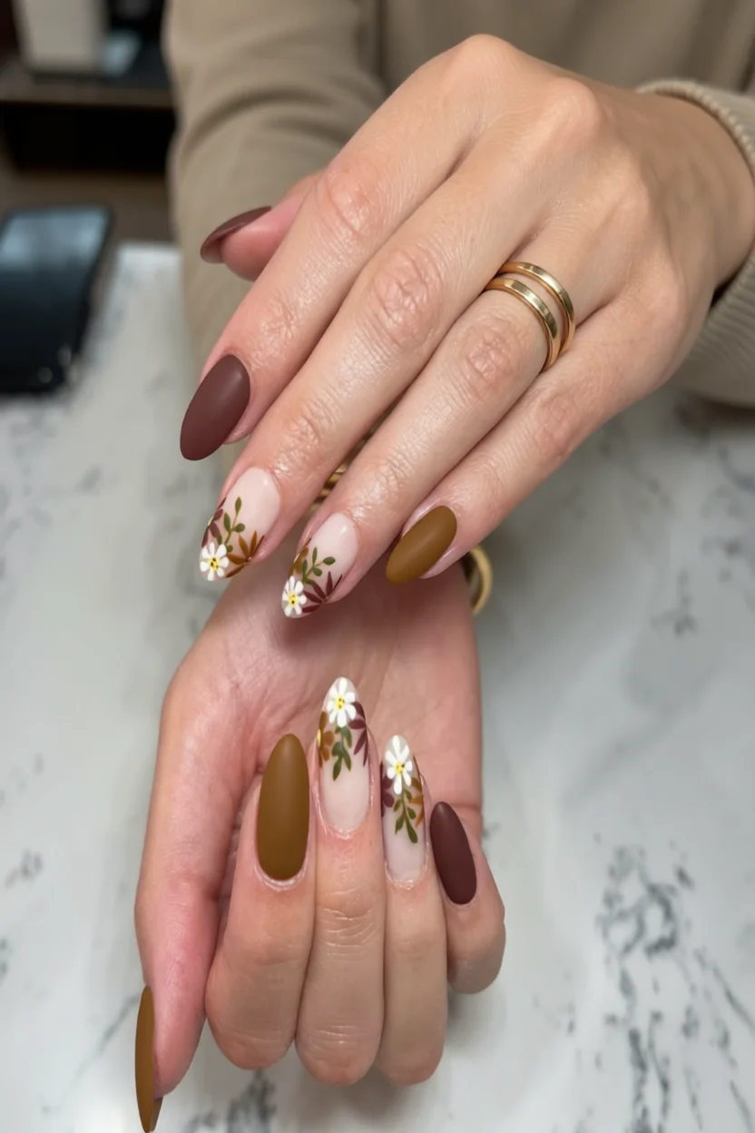 Uñas largas en forma de almendra con acabado mate en tonos chocolate, caramelo y flores blancas dibujadas a mano